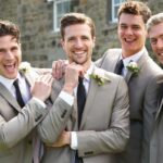 Quatro homens felizes, incluindo um noivo, padrinho e dois padrinhos, sorrindo para a câmera ao ar livre.