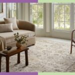 Sala de estar com sofá bege, tapete estampado, cadeira de madeira e três grandes janelas com vista para as árvores.