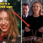 Pessoa com cabelos soltos e ondulados sorrindo, envolta em uma bandeira estampada, ao ar livre com vegetação desfocada ao fundo