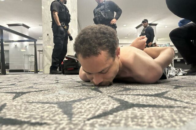 Um homem sem camisa está deitado de bruços sobre um tapete estampado com as mãos algemadas nas costas, enquanto três agentes de segurança em uniformes pretos estão de pé sobre ele.