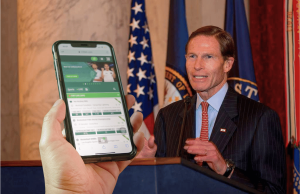 Blumenthal pressiona ligas esportivas por causa de laços de apostas e novas preocupações com integridade Senador dos EUA falando no pódio com bandeiras americanas enquanto o smartphone exibe a interface do aplicativo de apostas esportivas em primeiro plano, ilustrando preocupações com parcerias de jogos de azar e integridade esportiva. Blumenthal pressiona ligas esportivas por causa de laços de apostas e novas preocupações com integridade