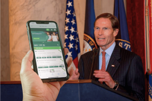 Blumenthal pressiona ligas esportivas por causa de laços de apostas Senador dos EUA falando no pódio com bandeiras americanas enquanto o smartphone exibe a interface do aplicativo de apostas esportivas em primeiro plano, ilustrando preocupações com parcerias de jogos de azar e integridade esportiva. Blumenthal pressiona ligas esportivas por causa de laços de apostas e novas preocupações com integridade