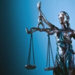 Uma estátua da Senhora Justiça segurando balanças em frente a um fundo azul.