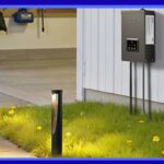 Um conjunto de transformador e luzes de caminho mostrado com uma caixa de transformador de metal escovado montada em uma casa e luzes de caminho iluminando um caminho de jardim à noite com pessoas reunidas ao fundo.