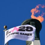 Uma bandeira branca dos Jogos Olímpicos LA28 balançando em frente a um caldeirão olímpico aceso contra um céu azul claro.