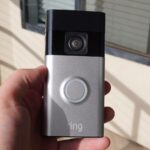 Uma campainha com câmera branca montada ao lado de uma porta branca, encostada em uma parede vermelha ousada com padrões geométricos.