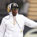 Deion Sanders, técnico do Colorado Buffaloes, com capuz branco, chapéu, óculos escuros e fones de ouvido dourados, em campo.