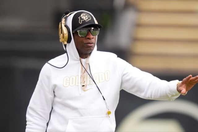 Deion Sanders, técnico do Colorado Buffaloes, com capuz branco, chapéu, óculos escuros e fones de ouvido dourados, em campo.