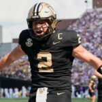 Um jogador de futebol americano de Vanderbilt, vestindo o número 2, com uniforme preto e dourado, comemora com os braços estendidos.