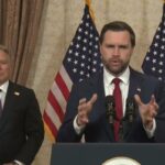 JD Vance falando em um pódio com duas bandeiras americanas atrás dele.