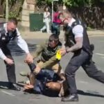 Os policiais chutam o suspeito na cabeça após aplicá-lo com um Taser. Eles foram ajudados por um transeunte (à direita)