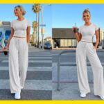 Uma mulher vestindo um top cinza claro de mangas curtas e calças largas combinando, segurando uma bebida e uma bolsa de ombro branca.