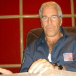 Jeffrey Epstein em uma camisa azul escura com um emblema da bandeira americana, sentado em uma cadeira.