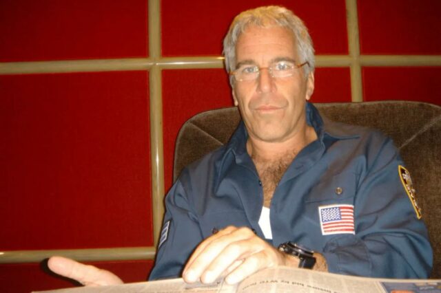 Jeffrey Epstein em uma camisa azul escura com um emblema da bandeira americana, sentado em uma cadeira.