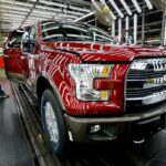 Um trabalhador inspeciona um caminhão Ford F-150 vermelho com a porta aberta em uma fábrica de montagem.
