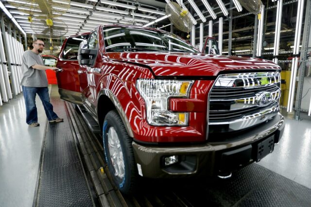 Um trabalhador inspeciona um caminhão Ford F-150 vermelho com a porta aberta em uma fábrica de montagem.
