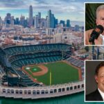 Um homem com cabelo curto e escuro e camisa de colarinho verde escuro gesticula com as mãos enquanto está sentado em uma cadeira branca.