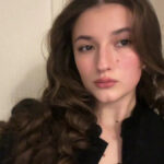 Uma garota morena está com seu boné e vestido preto, que apresenta um capuz de cetim amarelo pendurado em sua frente. Ela segura um ursinho de pelúcia marrom em ambas as mãos, que também usa um boné e um vestido em miniatura. A garota está parada em frente a uma escada.