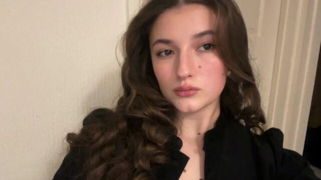 Uma garota morena está com seu boné e vestido preto, que apresenta um capuz de cetim amarelo pendurado em sua frente. Ela segura um ursinho de pelúcia marrom em ambas as mãos, que também usa um boné e um vestido em miniatura. A garota está parada em frente a uma escada.