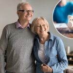 A mão de uma pessoa inserindo uma moeda em um cofrinho branco sobre uma mesa de madeira.