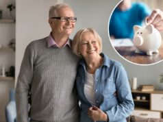 Há um novo ‘número mágico’ para as poupanças viverem ‘confortavelmente’ na aposentadoria – apenas metade dos americanos dizem que estão preparados A mão de uma pessoa inserindo uma moeda em um cofrinho branco sobre uma mesa de madeira.