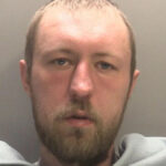 Um homem com um anoraque com capuz inspeciona um graveto encostado em uma cerca viva à noite