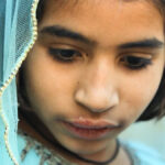 Um homem sentado em uma cadeira usando um estetoscópio, máscara facial e luvas cumprimenta uma mulher segurando seu filho. Os rostos da mulher e da criança estão obscurecidos.