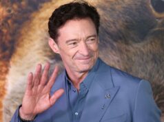 Hugh Jackman assume um novo papel em instituições de caridade inovadoras Hugh Jackman de terno azul, acenando, com uma cabeça de leão borrada ao fundo.