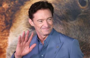 Hugh Jackman assume um novo papel em instituições de caridade inovadoras Hugh Jackman de terno azul, acenando, com uma cabeça de leão borrada ao fundo.