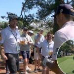 O jogador de golfe Jordan Spieth observa sua bola, mostrada com uma linha de trajetória amarela, voar pelo ar após acertá-la, enquanto os espectadores se alinham em ambos os lados do green.