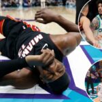 Um jogador de basquete de uniforme preto e vermelho segurando uma bola de basquete, saltando sobre outro jogador de uniforme branco e verde que caiu no chão.