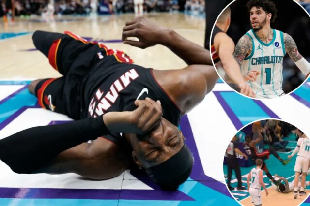 Um jogador de basquete de uniforme preto e vermelho segurando uma bola de basquete, saltando sobre outro jogador de uniforme branco e verde que caiu no chão.