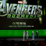 Robert Downey Jr. está no palco da Comic-Con fantasiado de Victor Doom e segura sua máscara voltada para o céu em um gesto triunfante