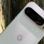 Uma mão segurando um smartphone Google Pixel azul com um ícone de alerta e ícones representando câmera, áudio e bugs.