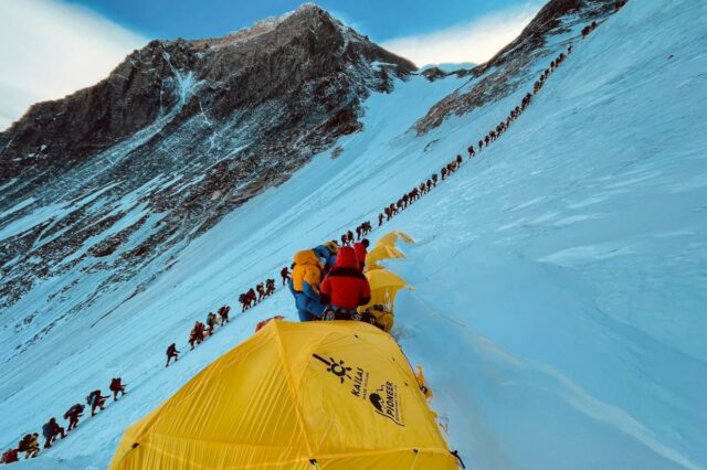 Alpinistas com equipamentos coloridos sobem o Monte Everest em uma longa fila, com tendas amarelas em primeiro plano.