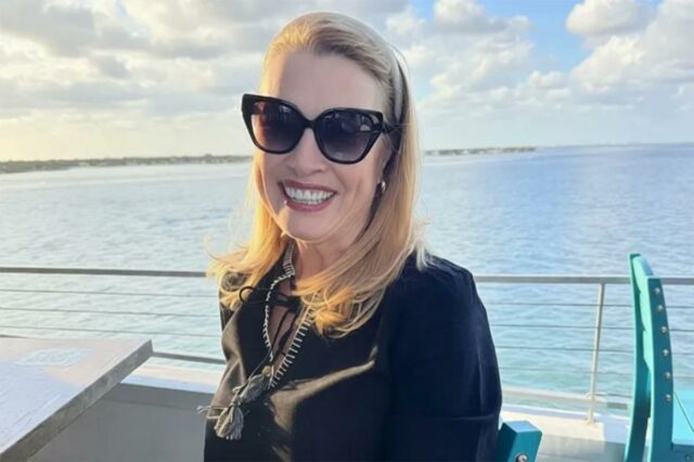 Eileen McGill Fox sorri para a câmera, usando óculos escuros e blusa preta, com um corpo d'água e o horizonte ao fundo.