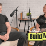 Artisan CEO, Jaspar Carmichael-Jack and Build Mode host Isabelle Johannessen sit facing each other in a podcast studio with microphones, mid-conversation, in front of a white brick wall; a small table with books and a plant sits between them, and a “TechCrunch Build Mode” logo overlay appears in the lower right corner.