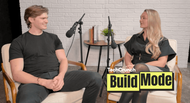 Artisan CEO, Jaspar Carmichael-Jack and Build Mode host Isabelle Johannessen sit facing each other in a podcast studio with microphones, mid-conversation, in front of a white brick wall; a small table with books and a plant sits between them, and a “TechCrunch Build Mode” logo overlay appears in the lower right corner.