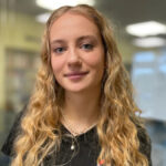 Daisy tem cabelos castanhos longos e lisos e olhos escuros. Ela está vestindo uma camiseta preta e tem dois colares de prata no pescoço.