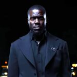 A man in a black suit standing in front of a dark backdrop