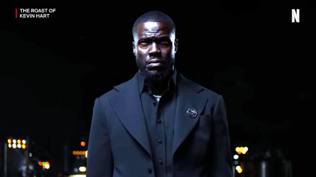 A man in a black suit standing in front of a dark backdrop