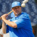 Um jogador de beisebol com boné e camisa azuis segura um taco com uma bola de beisebol acima do ombro.