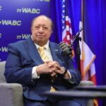 Um homem de terno azul e gravata amarela listrada sentado em uma poltrona cinza, em frente a um fundo azul com o