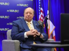 O bilionário John Catsimatidis comemora seu primeiro fim de semana como gerente de uma liga secundária de beisebol Um homem de terno azul e gravata amarela listrada sentado em uma poltrona cinza, em frente a um fundo azul com o