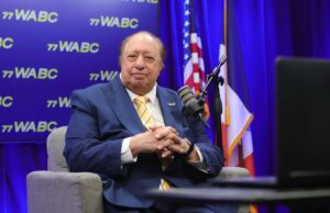 O bilionário John Catsimatidis comemora seu primeiro fim de semana como gerente de uma liga secundária de beisebol Um homem de terno azul e gravata amarela listrada sentado em uma poltrona cinza, em frente a um fundo azul com o