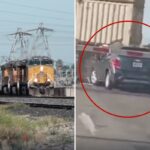 Um SUV de cor escura está preso nos trilhos de uma ferrovia enquanto um trem se aproxima, seu pára-choque traseiro batendo na lateral de um vagão de carga.