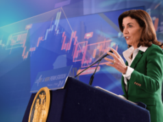 O governador Hochul proíbe negociações de mercado de previsão privilegiada para trabalhadores do estado de Nova York A governadora de Nova York, Kathy Hochul, fala no pódio enquanto o gráfico do mercado de ações ilustra a ordem executiva que proíbe o uso de informações privilegiadas nos mercados de previsão.