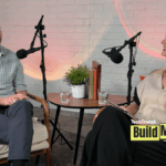 A podcast-style interview setup shows a man and a woman seated across from each other in armchairs, speaking into microphones. They sit beside a small round table with water glasses, books, and a potted plant, against a white brick wall backdrop. A “TechCrunch Build Mode” logo appears in the lower corner.