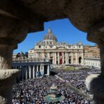Ampla vista da Praça de São Pedro repleta de multidão durante a missa de Páscoa, emoldurada por uma abertura arquitetônica.