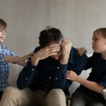 Um pai exausto está sentado em um sofá com o rosto entre as mãos, enquanto seus dois filhos pequenos, um menino e uma menina, ficam um de cada lado, tocando-o.